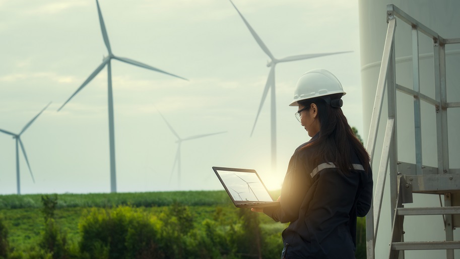 Female Engineer Wearing Hard Hat Standing With Digital Tablet Against Wind Turbine.